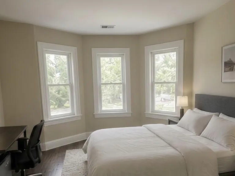 Furnished bedroom with a bay of three windows, queen bed, bedside tables, and a desk workspace with TV.