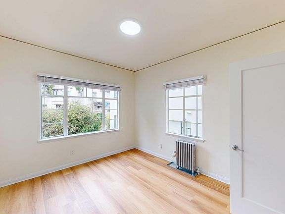 Bright, empty room with hardwood flooring, two windows, a radiator, and natural light fills the space