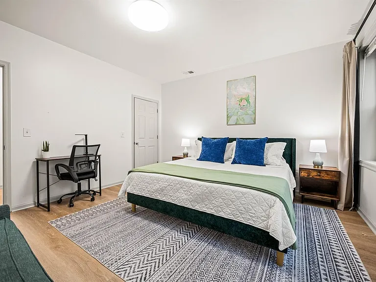 Spacious furnished bedroom with a queen bed, nightstands, and a dedicated desk area, plus large windows with curtains.