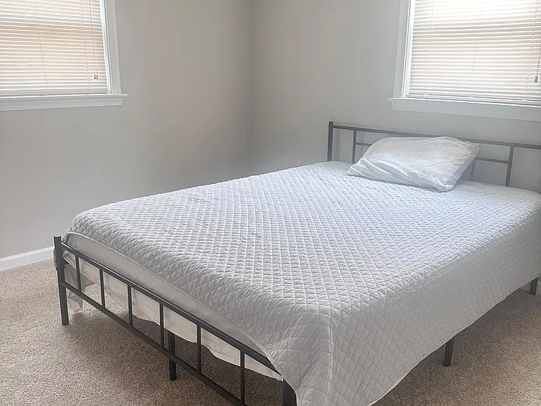 Furnished bedroom with a full-size bed, soft carpet, and two windows with blinds for flexible light and privacy.