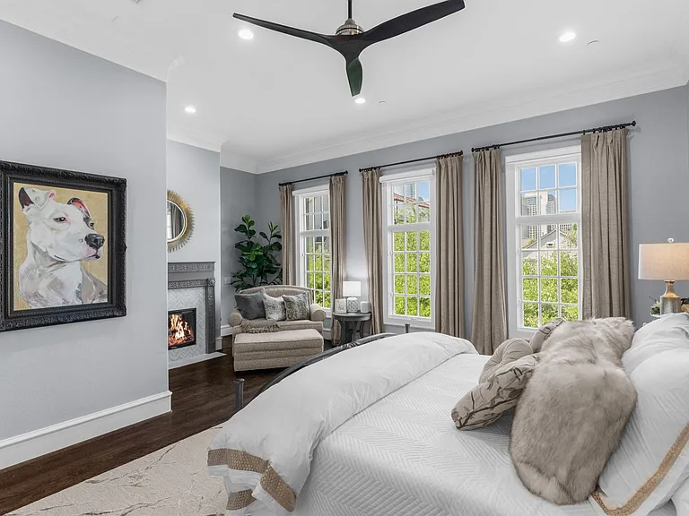 The 3rd-floor is dedicated to the primary suite with tall windows, soft gray walls, dark wood floors, an indoor fireplace, and a neutral-toned bed styled with layered pillows and textured bedding.