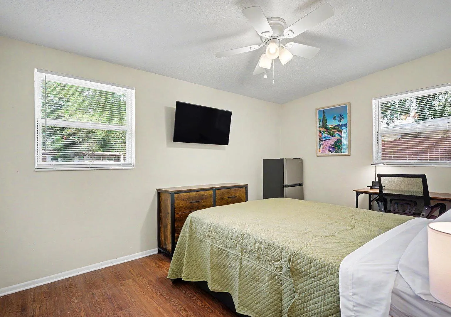Large bedroom w/2 windows, ceiling fan, wall-mounted 40" TV, 6-drawer dresser, work desk w/lamp, and an in-room mini fridge.