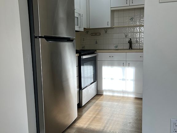 Kitchen with brand new appliances, refrigerator, electric oven, and dishwasher. Microwave above stove. There is ample cabinet space, and an open shelving unit for pantry items. Wall behind sink is tiled with floral pattern.