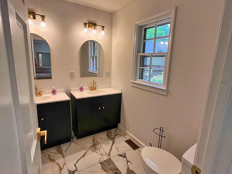 Updated bathroom with dual-sink vanity, sleek gold fixtures, large tile flooring, and a window for natural light.
