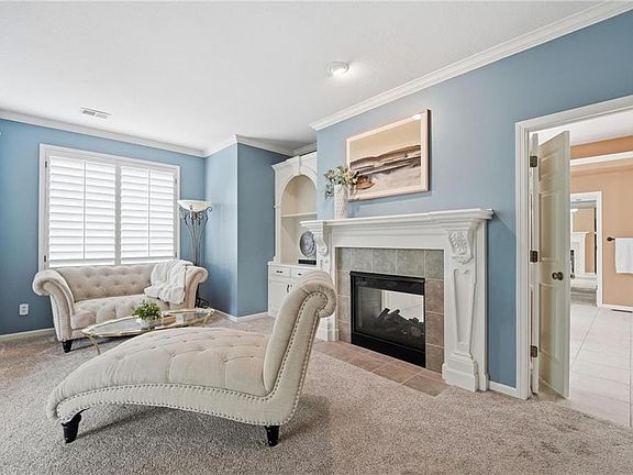Carpeted room featuring a fireplace with a decorative mantel and stone surround, built-in cabinetry, and white plantation shutters on the window