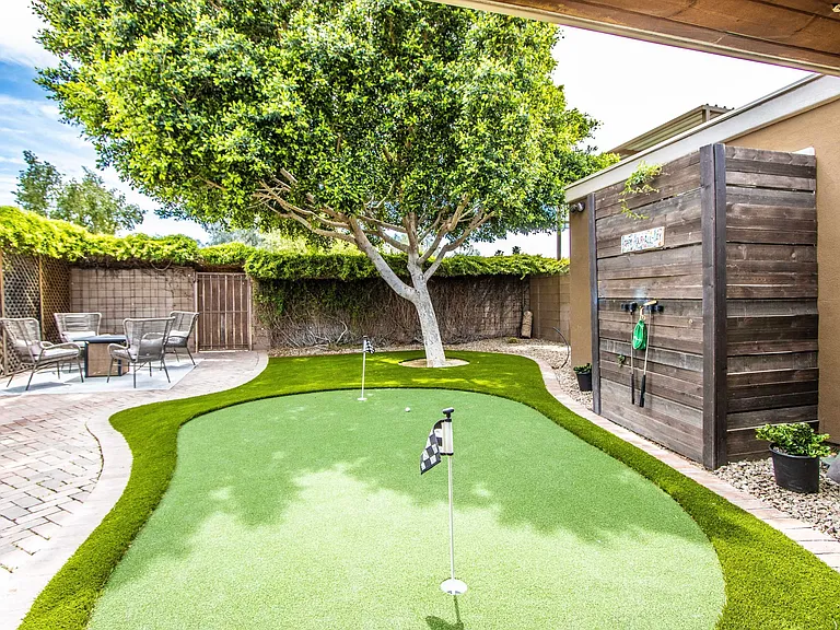 Beautiful putting green at Casa Del Sol, Tempe rental home. Showcases a custom artificial turf green with golf flags, a rustic wood feature wall with putter storage, a shade tree, and a patio fire pit set. Perfect outdoor space for a Tempe, AZ stay.