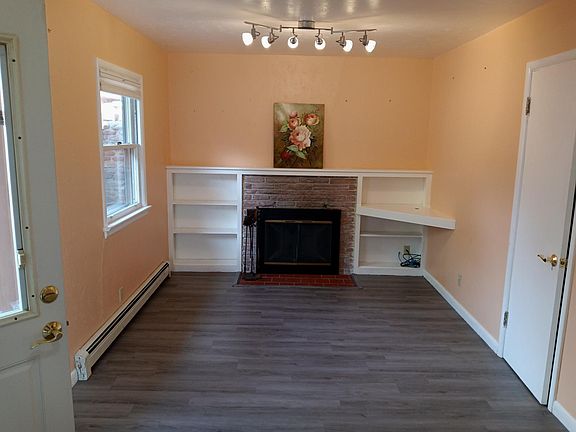 The living room area, showing the fire place with a screen and glass doors, the window with blinds, the entrance door to left, the coat closet to right, and the dimmable spotlights on the ceiling.