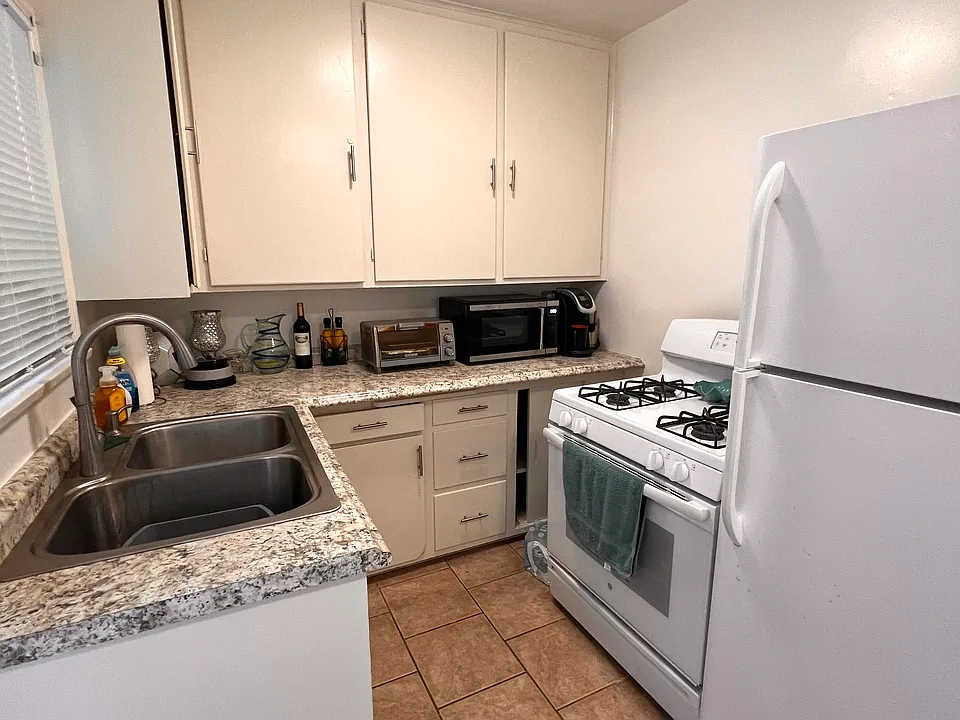 Kitchen with large refrigerator and gas stove. Large window above sink and another large window at entrance.