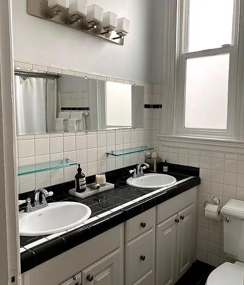 Bathroom with 2 sinks, toilet, and tub shower with hotel double rod for spacious bathing experience. Lots of natural light.