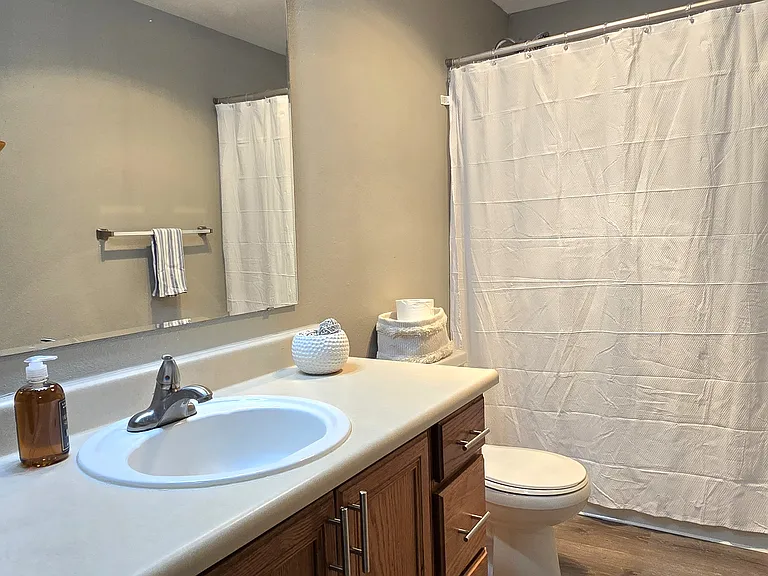 Clean, well-lit bathroom featuring a spacious vanity with ample counter space, warm wood cabinetry, and a full tub with shower. Neutral finishes create a bright, inviting space for everyday use.