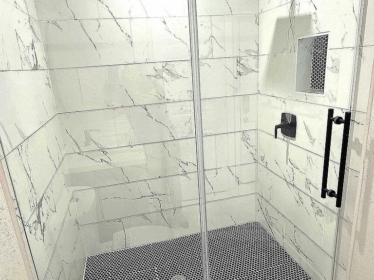 Modern bathroom with a sleek glass-enclosed shower featuring white tiles, a rainfall showerhead, and a stylish black hexagon-tiled floor. The space is bright and contemporary, providing a luxurious feel.