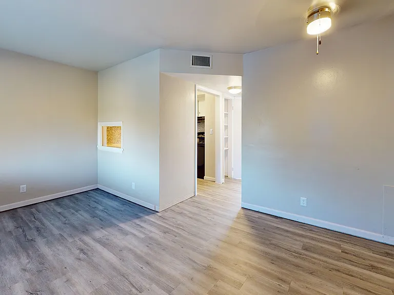 Bright, modern apartment room with stylish wooden flooring and soft lighting, featuring an open doorway leading to a cozy kitchen.