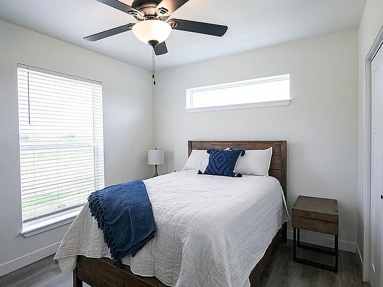 Charming bedroom with modern ceiling fan, cozy wooden bed, and ample natural light from large windows.