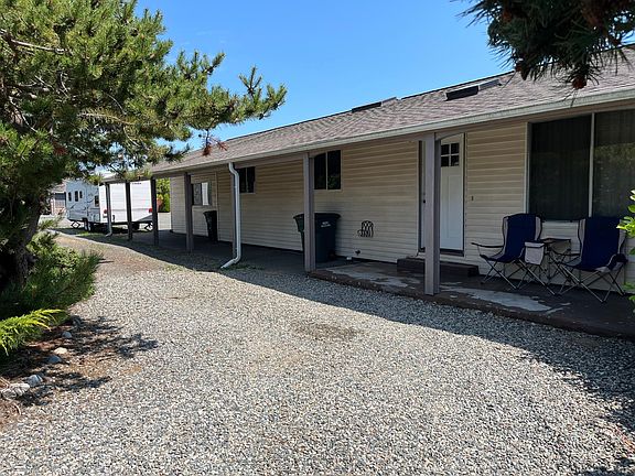 Front of the house from Quinault showing the gravel circular driveway (providing extra parking) in the foreground, covered walkway from garage past two bedrooms and the living room window o the right.