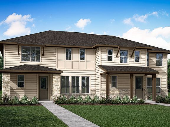 A two-story beige residential building with a dark roof stands in a grassy area surrounded by trees