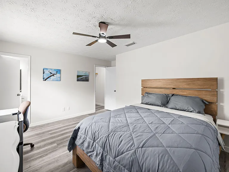Furnished bedroom with a queen bed, ceiling fan, and dedicated desk space, plus easy access to the closet and hallway.