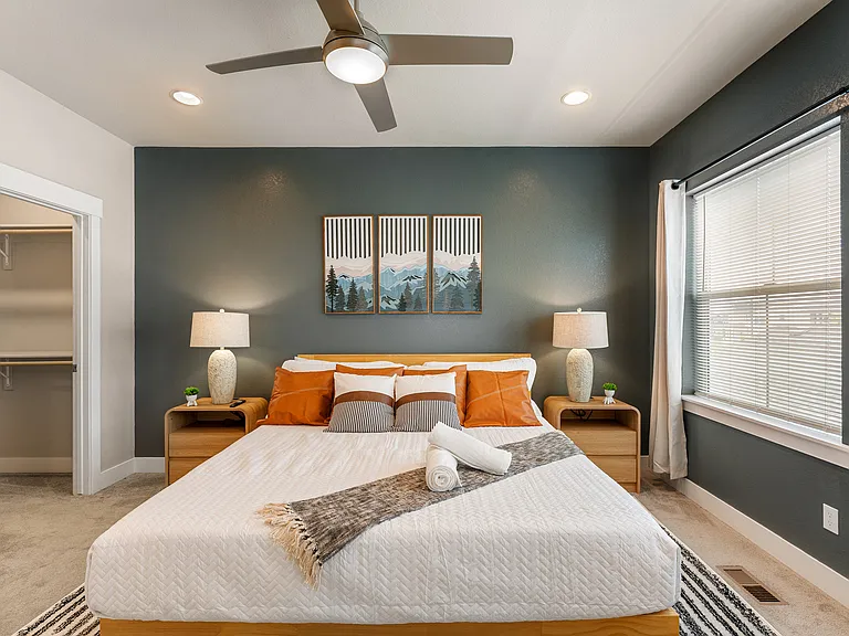 Stylish bedroom with a cozy king-sized bed dressed in white and burnt orange accents. Two bedside tables with elegant lamps flank the bed. The room features a ceiling fan, a walk-in closet, and large windows for plenty of natural light.