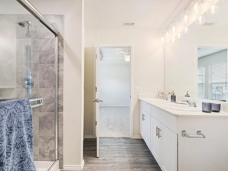 Contemporary bathroom featuring a glass-enclosed shower with grey tile surround, a double vanity with white cabinetry, a large mirror, and wood-finish flooring