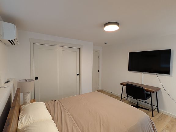 Shows the queen size bed, dedicated workspace, smart TV, wall mounted mini split, closet, side table with lamp.