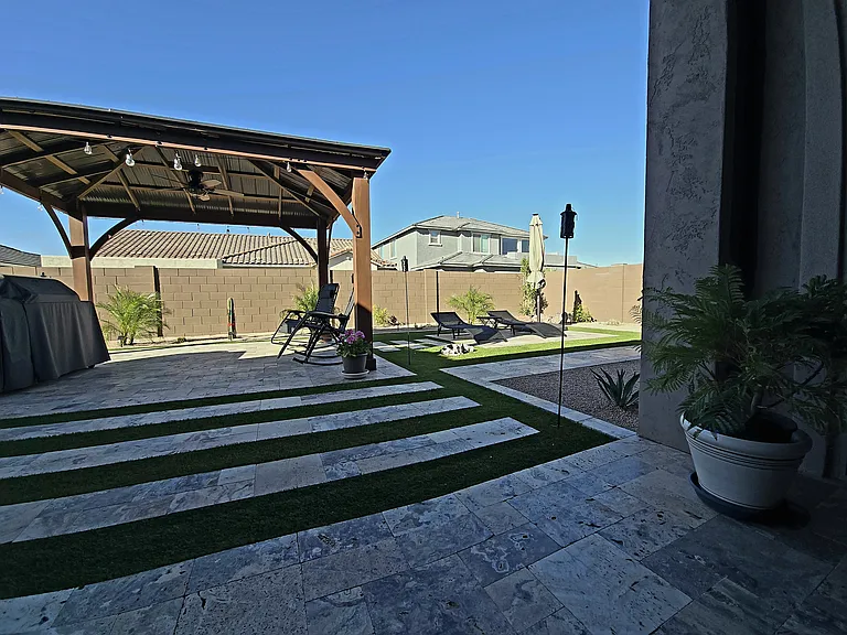 This inviting backyard features a pergola with seasonal decor over a patio dining set, plus a grill and lounge chairs for sunbathing. Travertine tiles with grass inlays, potted plants, and a privacy wall create a stylish space for relaxing or entertaining