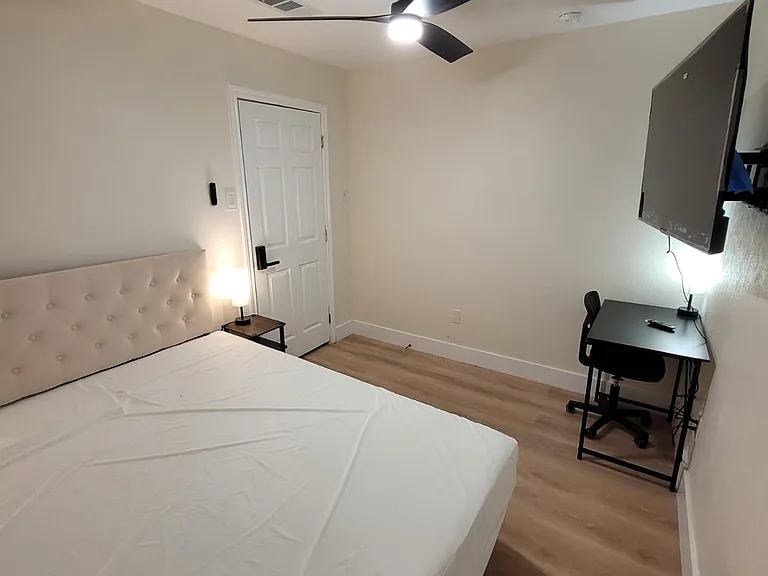 Private bedroom with a bed, nightstand lighting, ceiling fan, and a dedicated desk area with wall-mounted TV.