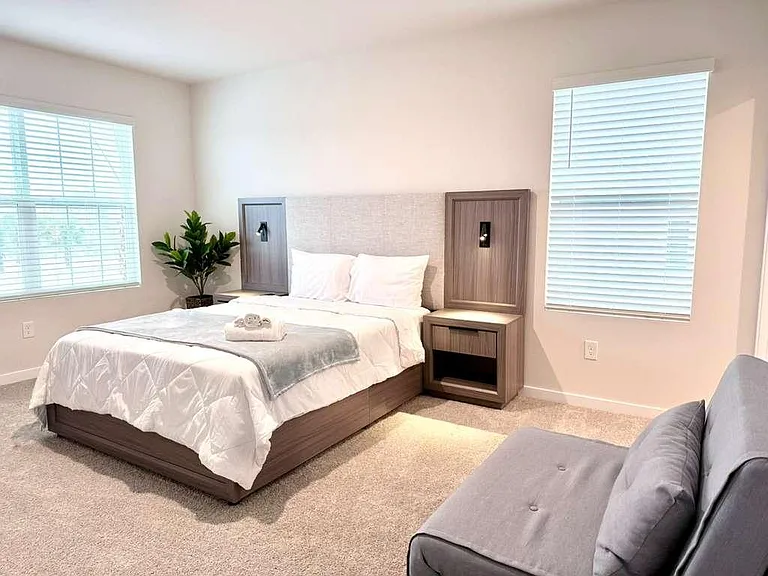 Spacious bedroom with a queen bed, upholstered headboard, and matching nightstands, plus two windows and soft carpet.