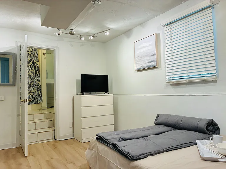 Furnished bedroom with a comfortable bed, dresser with TV, window blinds, and quick access to an adjacent bathroom.