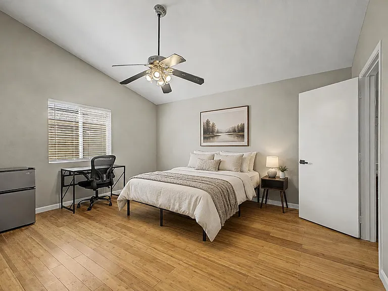 Spacious furnished bedroom with hardwood floors, ceiling fan, work desk, and a mini fridge for added convenience.