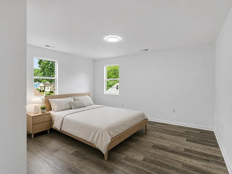 Spacious bedroom with wood-look flooring, overhead lighting, and two windows offering neighborhood and treetop views.