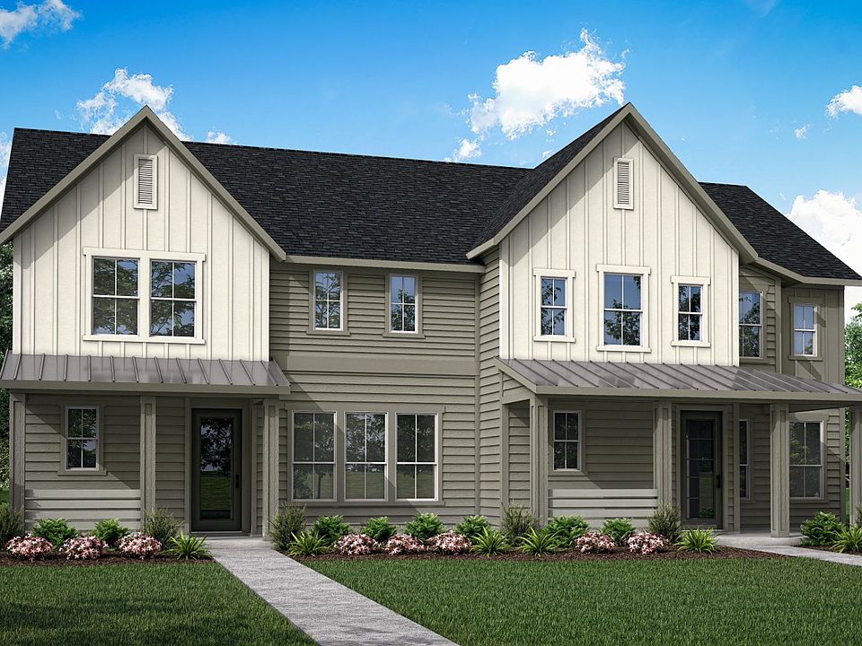 A two-story beige house with a black roof, surrounded by lush greenery and a grassy lawn in the fore