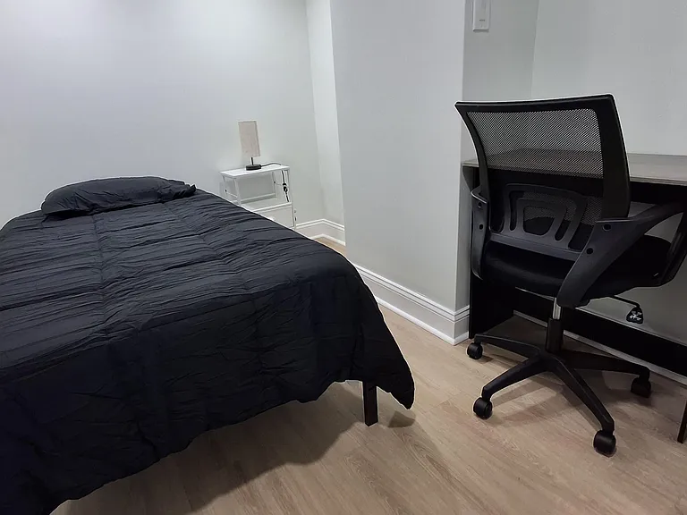 Private furnished bedroom with a full bed, bedside table, and dedicated desk setup for work or study.