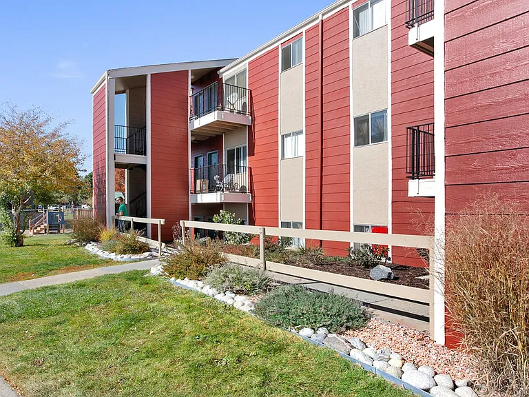Charming red apartment building with balconies, surrounded by lush landscaping and a peaceful walkway.