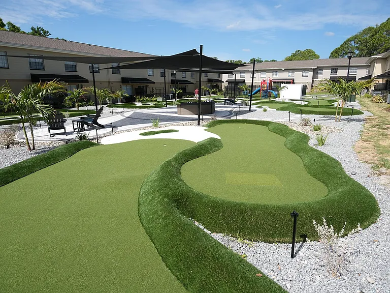 Charming apartment complex courtyard featuring a mini-golf course, lush landscaping, and outdoor seating under stylish canopies.