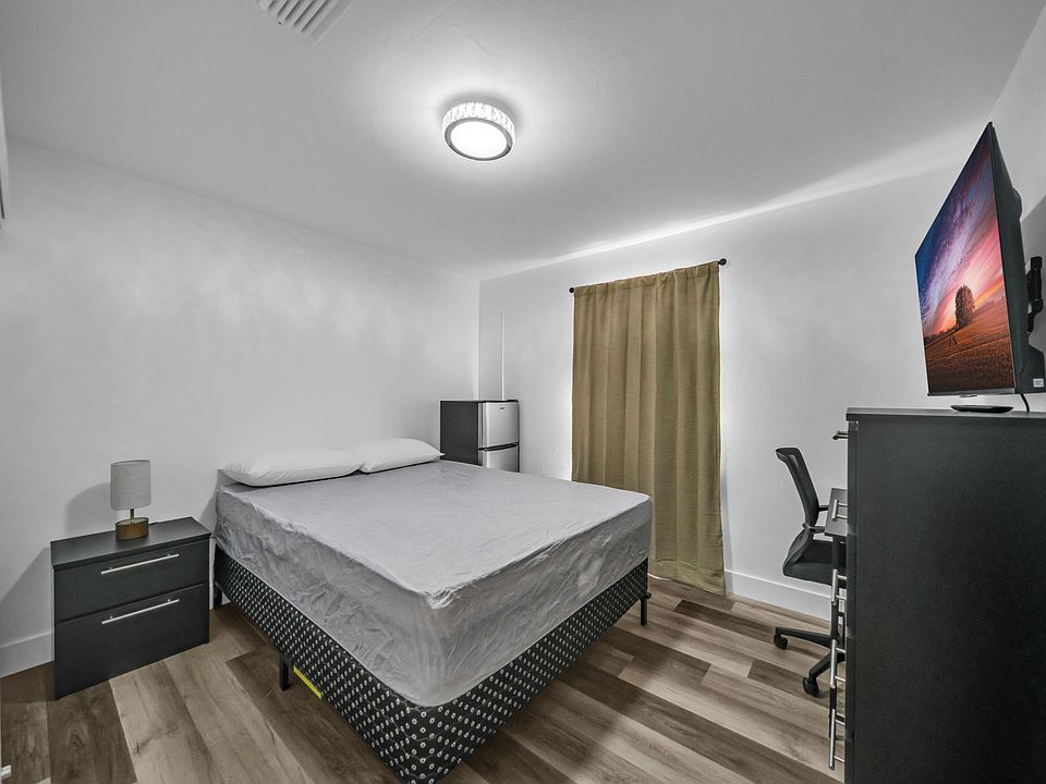 Furnished bedroom with a full bed, nightstand, mini fridge, and a work desk, plus a window with privacy curtains.