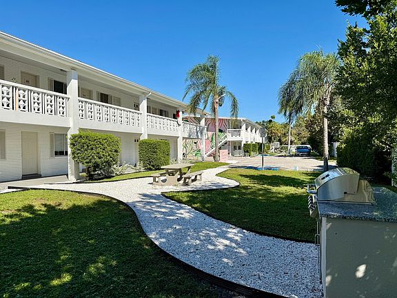 Charming two-story apartment complex with lush greenery, palm trees, and a modern outdoor BBQ area, perfect for relaxation.