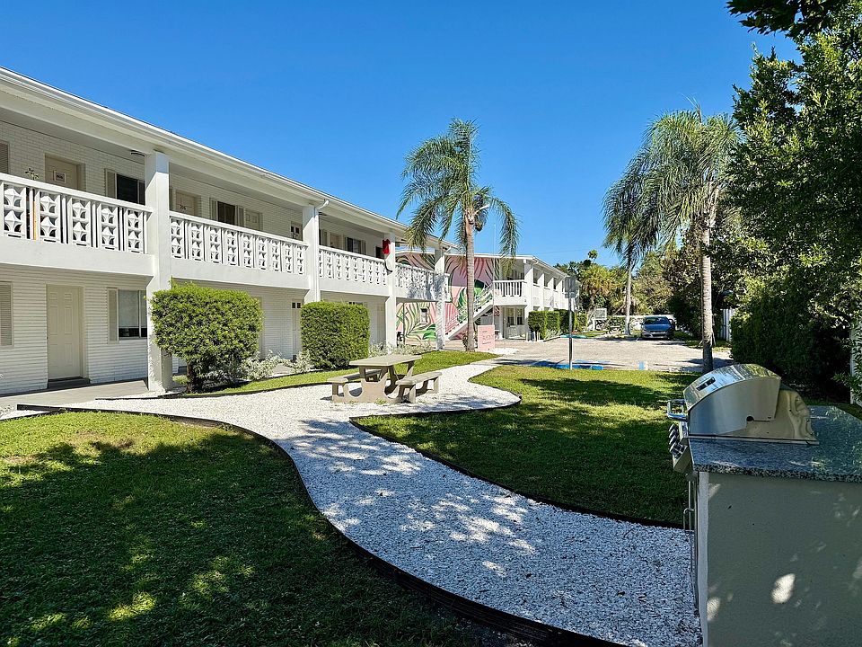 Charming two-story apartment complex with lush greenery, palm trees, and a modern outdoor BBQ area, perfect for relaxation.