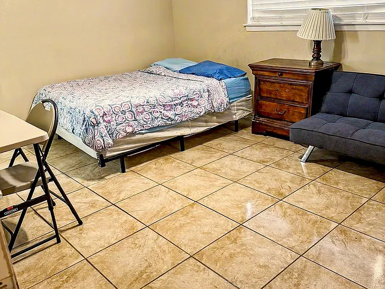 Furnished bedroom with tile flooring, a bed, nightstand with lamp, and a seating area near the window with blinds.