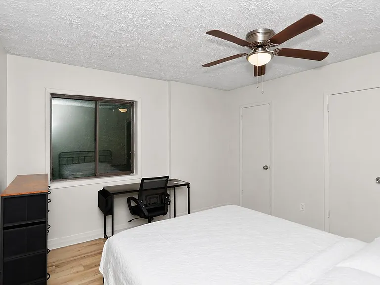 Furnished bedroom with a ceiling fan, large window, and a dedicated desk/work area plus storage for everyday essentials.