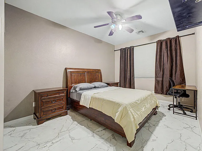 Furnished bedroom with a full bed, nightstand, and desk, plus a large window with curtains and a ceiling fan light.