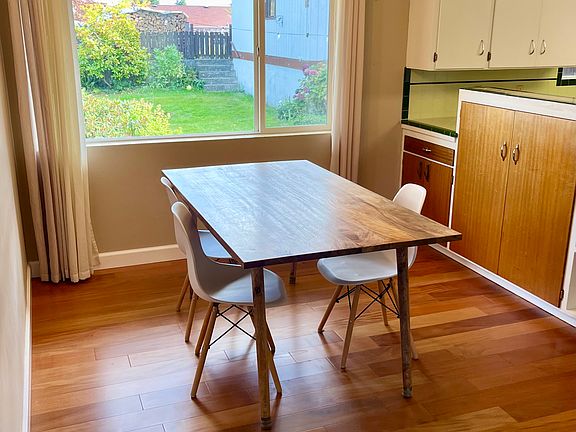 Dining area with wood floors, modern chandelier, and backyard views through large sunny window.