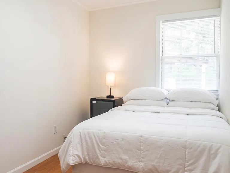 Furnished bedroom with a full-size bed, nightstand lighting, and a large window with blinds for comfortable daily living.