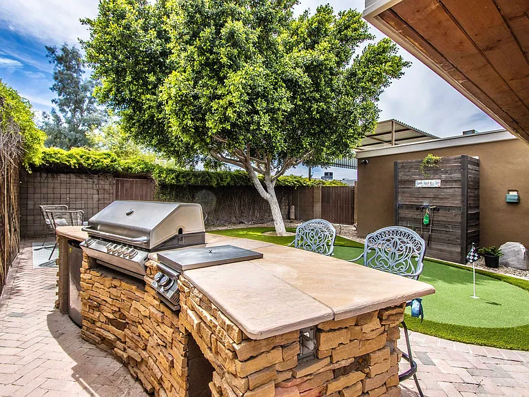 Premium BBQ area at Casa Del Sol, Tempe rental home. Showcases a custom stone grilling island with a built-in stainless steel grill, bar seating, brick pavers, and a custom putting green with a lush shade tree. Premier outdoor Tempe, AZ stay.