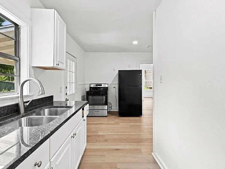 Efficient kitchen space featuring white shaker cabinetry, dark stone countertops, a dual basin stainless steel sink with a gooseneck faucet, light wood-finish flooring, and white walls