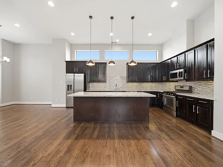 Beautifully designed kitchen featuring dark wood cabinetry, sleek stainless steel appliances, and a large center island ideal for hosting. Soaring ceilings, stylish pendant lighting, and continuous hardwood flooring create a bright, open space.