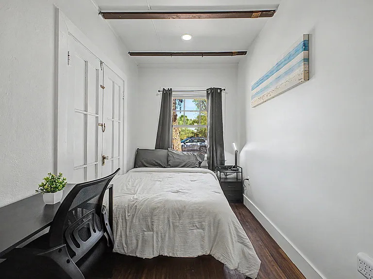 Private bedroom with a full bed, bedside table, and desk setup, plus a window with curtains for natural light.