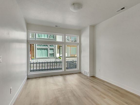 A modern, empty room with light wood flooring, white walls, and large windows facing a fenced outdoo