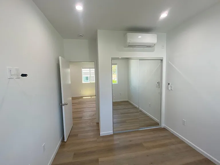 Bright bedroom with mirrored closet doors, mini air conditioning, wood flooring, and clean white walls.