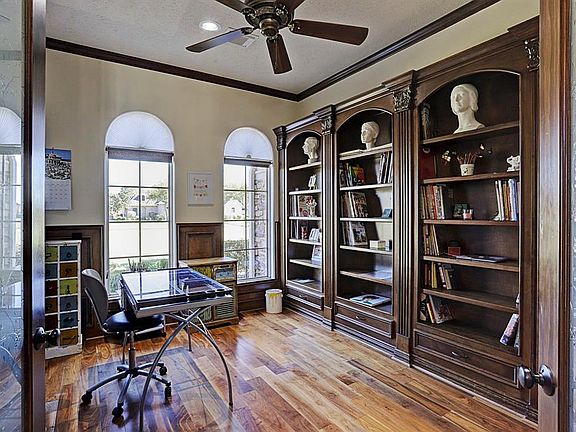 The study has rich hardwood floors a wall of custom built bookshelves, wood wainscoting around the room, double French doors, crown molding, and tall ceiling with fan.