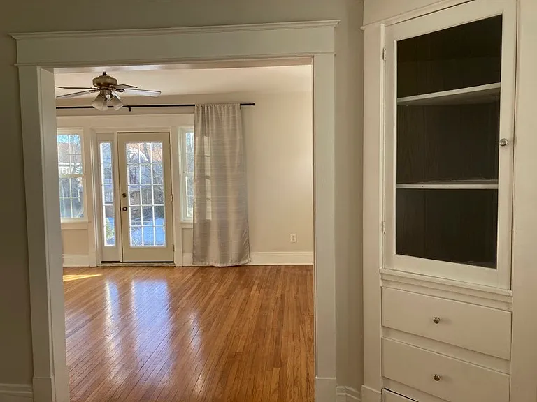 Living room, as viewed from dining room. Door to front porch, ceiling fan, and window treatments (can be included). Dining room built-in cabinets also visible.