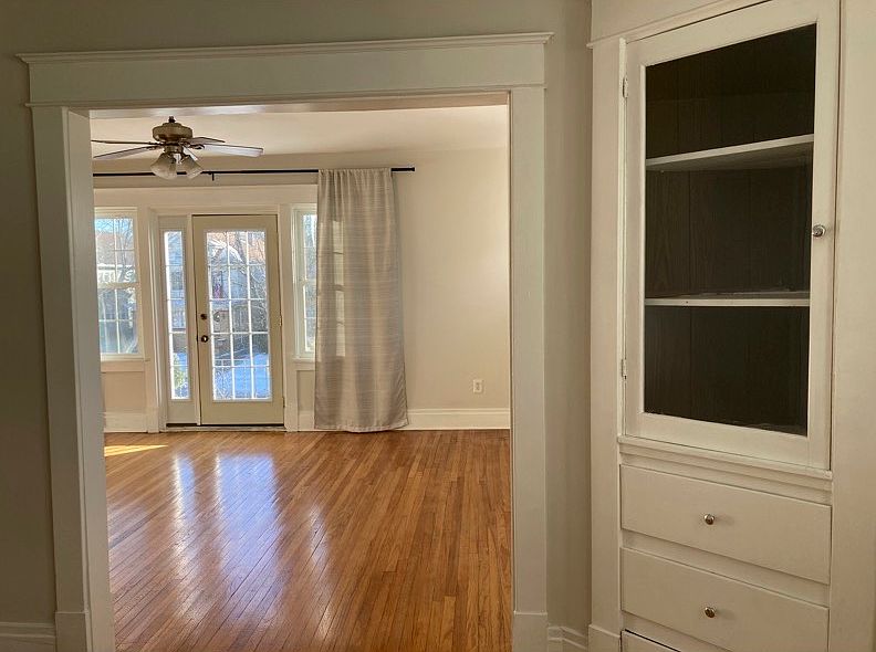 Living room, as viewed from dining room. Door to front porch, ceiling fan, and window treatments (can be included). Dining room built-in cabinets also visible.
